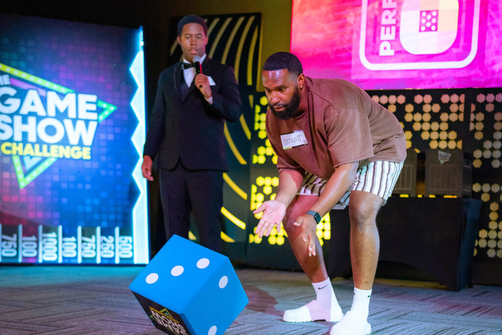 Host with a suit, tie and microphone provides commentary while a guest rolls a giant dice. 