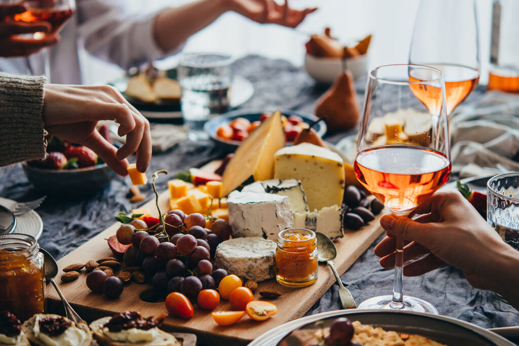 Overhead view of a shared charcuterie and cheese board with grapes, nuts, and glasses of rosé wine as friends reach in together