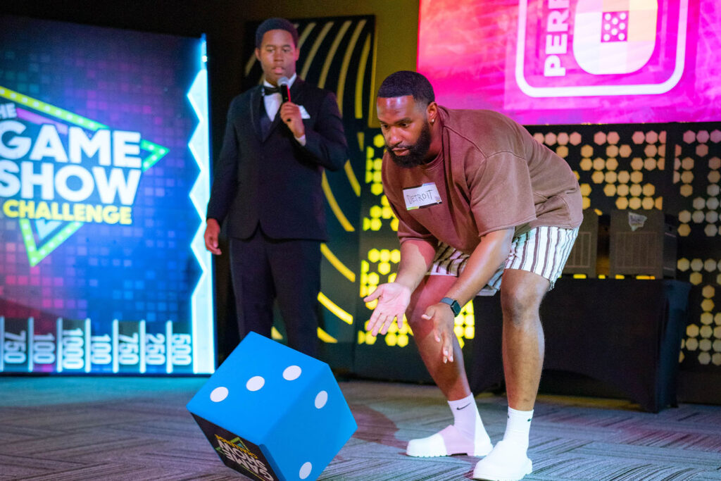 A Game Show Challenge contestant tossing an oversized foam die on stage while a tuxedoed host holds the microphone, in front of the Game Show Challenge neon logo